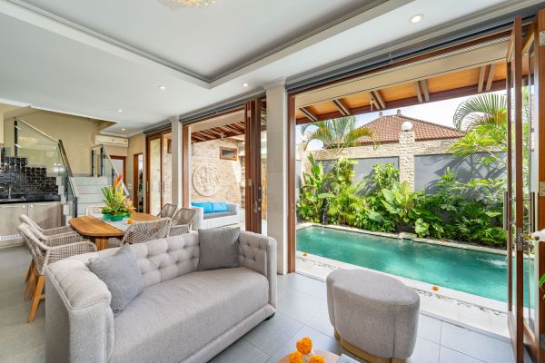 Modern Balinese villa interior with open-plan lounge, gray sofa, dining table, and folding doors to turquoise pool amid tropical gardens. - Bali Villas